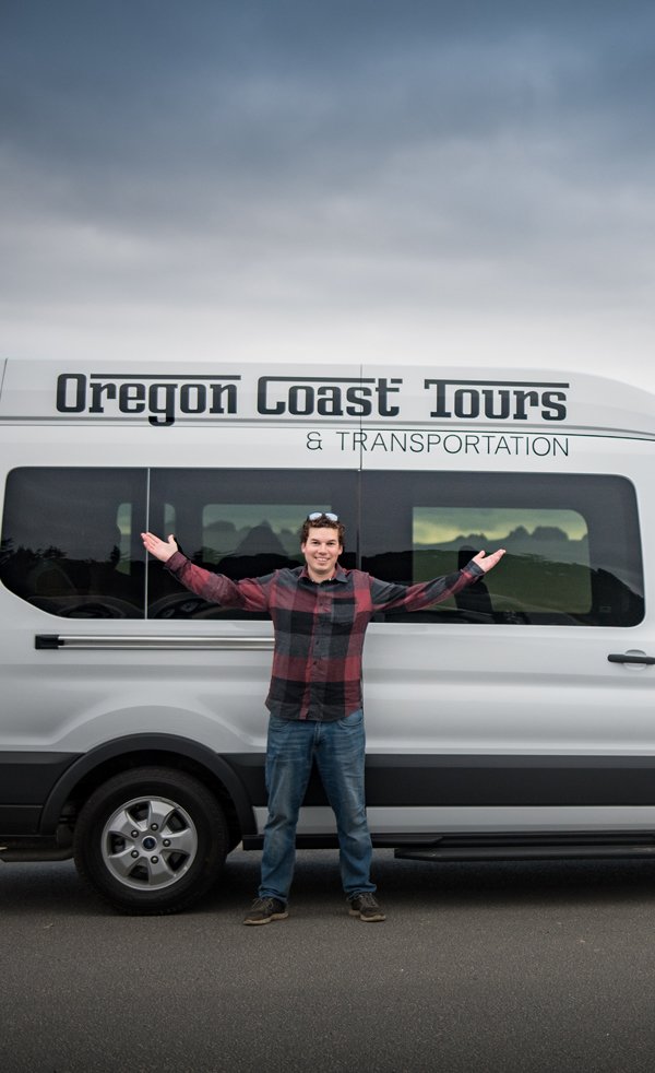 Owner of Oregon coast tours, Charlie, standing in front of his van with his arms stretched out near the beach on the oregon coast
